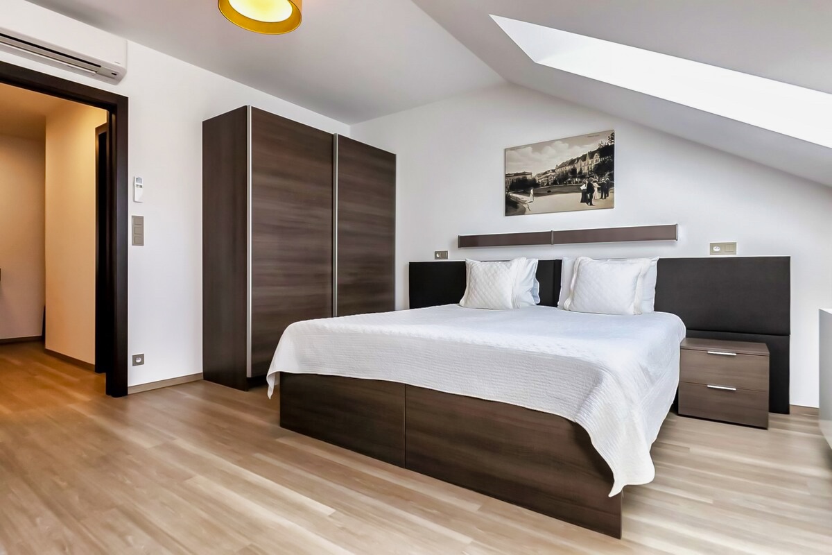 Discover the charm of Aparthotel Vinohradský dům Prague Studio Attic, featuring a serene bedroom with elegant wooden floors and a pristine white bed